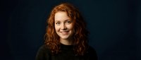 Young redhead woman with curly hair smiling warmly at camera wearing dark green sweater against black background for professional headshot photography.