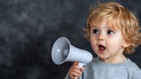 Excited toddler with curly blonde hair holding megaphone against dark background, expressing surprise and joy. Concept of children communication and voice.