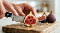 Fresh fig cut in half on wooden cutting board with knife in hand, showing red interior and seeds, food preparation and cooking concept.