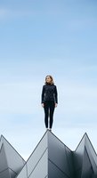 Young woman with shoulder-length blonde hair, fair complexion, wearing dark patterned sweater and black leggings, standing confidently on geometric architectural structure against clear blue sky for motivational and leadership concepts.