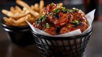 Crispy fried chicken wings glazed in sweet and spicy sauce garnished with fresh herbs served in basket with golden french fries for restaurant menu.