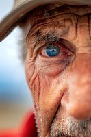Close-up portrait of senior man with intense blue eyes, weathered skin, and gray stubble wearing cowboy hat, capturing authentic aging and wisdom in natural light.
