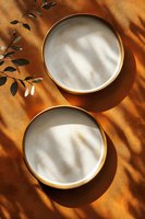 Empty white ceramic plates with gold rim on orange background with natural leaf shadows and sunlight creating minimalist composition for food styling.