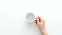 Hand holding an empty white ceramic coffee cup from above on a clean white background, minimalist flat lay style.