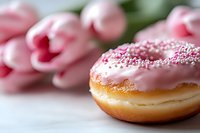 Pink glazed donut with sprinkles on white surface, fresh pink tulips in soft focus background creating romantic spring mood for food photography and social media.