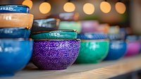 Colorful ceramic bowls with ornate patterns arranged in row on shelf, featuring vibrant purple, blue, and turquoise colors with bokeh light background.