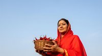 Middle-aged woman of South Asian ethnicity wearing traditional red headscarf and clothing, holding wicker basket of red chili peppers against clear blue sky. Suitable for agriculture, farming, harvest, and cultural lifestyle content.