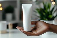 African American female hand holding blank white cosmetic tube against blurred bathroom background with bokeh lights and plants. Mockup for skincare product.