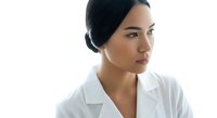 Young woman with dark hair in a bun, olive complexion, wearing white lab coat, looking thoughtfully to the side against white background. Healthcare professional portrait.