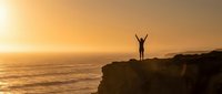 Person with raised arms celebrating success on cliff edge during golden sunset over ocean waves creating inspirational moment of achievement and freedom.