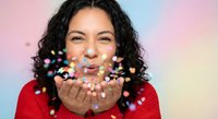 Young Hispanic woman in red sweater blowing colorful confetti from hands against pastel gradient background, celebrating joy and festive moments.