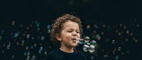 Young mixed-race boy with curly hair blowing soap bubbles against dark bokeh background, expressing joy and childhood wonder in studio portrait.
