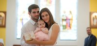 Young Caucasian couple with their infant daughter dressed in pink at a religious ceremony in a church with stained glass windows and yellow walls.