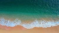 Aerial view of turquoise ocean waves gently rolling onto sandy beach, creating white foam patterns along shoreline. Natural summer seascape from above shows pristine coastline.