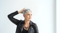 Mature woman with short gray hair wearing black leather jacket and black gloves, hand to head in thoughtful pose against white background for portrait and lifestyle use.