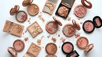 Luxury makeup collection featuring bronze and rose gold cosmetics including blush, bronzer, eyeshadow palettes scattered on white background.