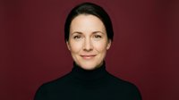 Professional Caucasian businesswoman in black turtleneck smiling confidently against burgundy background for corporate headshot and marketing materials.