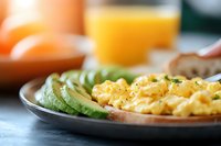 Fluffy scrambled eggs garnished with herbs served alongside fresh sliced avocado on gray plate, orange juice glass in background, morning breakfast setting.