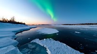 Spectacular northern lights dance over frozen lake landscape with snow-covered shores and cracked ice surface during winter twilight at arctic circle.
