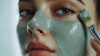 Young woman applying green clay facial mask with cosmetic brush during skincare routine and beauty treatment at spa salon. Close up view of fresh skin.