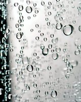 Clear water drops on glass surface create abstract pattern with varied sizes of spherical droplets against blurred gray background. Macro photography.