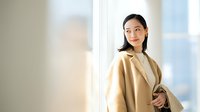 Professional Asian businesswoman in beige coat smiling confidently in modern office environment with natural lighting for corporate presentations.