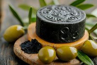 Black activated charcoal soap bar on wooden holder with green olives and olive leaves, natural skincare product for detoxifying facial cleansing.