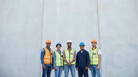 Diverse construction team of five workers wearing safety helmets and high-visibility vests standing against concrete wall at building site.