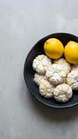 Fresh lemon crinkle cookies dusted with powdered sugar served in dark bowl with whole lemons on gray concrete surface for baking and dessert concepts.