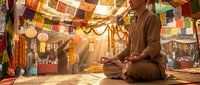 Young Asian monk in brown robes meditating peacefully in temple courtyard with colorful prayer flags and warm golden sunlight streaming through.
