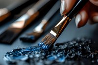Artist hand holding paintbrush applying dark blue paint on textured canvas surface. Creative art supplies and brushes blurred in background for artistic projects.