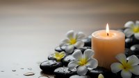 Spa wellness scene with burning candle, black massage stones and white plumeria flowers on wooden surface for relaxation and meditation therapy.