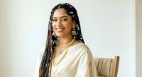 Young woman with dark skin tone and long braided hair with beaded accessories wearing cream-colored top, seated indoors for portrait use.