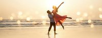 Romantic couple dancing on beach at sunset with golden light and ocean waves, man lifting woman in flowing red dress during magical golden hour moment.