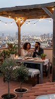 Young mixed-race couple enjoying romantic dinner on rooftop terrace with string lights and city skyline view at dusk for outdoor dining and date night.