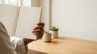 Person with dark skin tone holding open spiral notebook at wooden desk with potted plant and cup, workspace organization and planning concept.