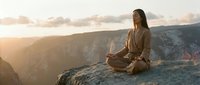 Young woman meditating in lotus position on mountain cliff edge during golden hour sunset with dramatic canyon landscape and warm sunlight.