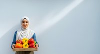 Woman wearing white hijab and blue cardigan holding wooden crate of fresh colorful bell peppers against white wall with natural light for food, agriculture, and healthy eating concepts.
