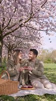 Young couple enjoying spring picnic under blooming cherry blossom trees, sharing dessert and romantic moments outdoors during springtime celebration and leisure activity.
