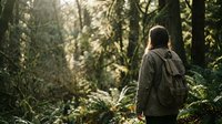 Hiker with backpack walking through dense forest trail surrounded by tall trees and natural sunlight filtering through canopy.