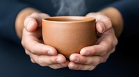 Gentle hands holding warm clay cup with rising steam, showcasing mindful moment and comfort. Close-up on terracotta ceramic mug against dark background.