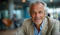 Confident senior Caucasian businessman with white hair and friendly smile wearing beige suit jacket and blue shirt against blurred office background.