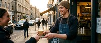 A male vendor with a beard and brown hat hands a newspaper to a female customer on a busy urban street lined with historic buildings and storefronts during daytime.