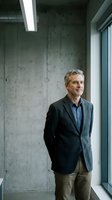 Confident mature Caucasian businessman in suit jacket standing by office window against concrete wall, looking thoughtfully into distance in modern workspace.