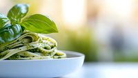 Fresh pasta with homemade basil pesto sauce served in white ceramic bowl against soft blurred background, garnished with fresh basil leaves, macro food photography.