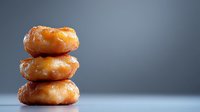 Stack of three glazed donuts on minimalist gray background, showcasing golden-brown fried dough with shiny sugar coating for food photography or bakery marketing.
