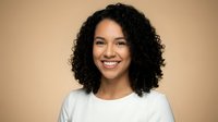 Young mixed-race woman with curly hair smiling warmly at camera wearing white casual top against neutral beige background for portrait photography.