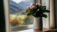Pink roses in textured ceramic vase on windowsill against mountain landscape background, soft natural lighting creates cozy home atmosphere.
