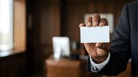 Businessman holding blank white business card in hand, professional corporate environment with office background, networking and contact information concept.