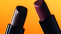 Dark burgundy lipsticks in black tubes against vibrant orange background with dramatic lighting and reflective surface, close-up macro shot showing luxurious cosmetic texture.